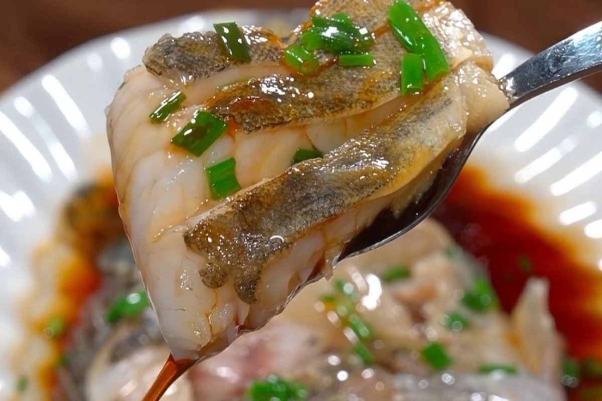 Close-up of steamed fish flesh — chopsticks lifting a pearlescent piece from the bone, translucent edges catching light, soy-ginger oil glistening