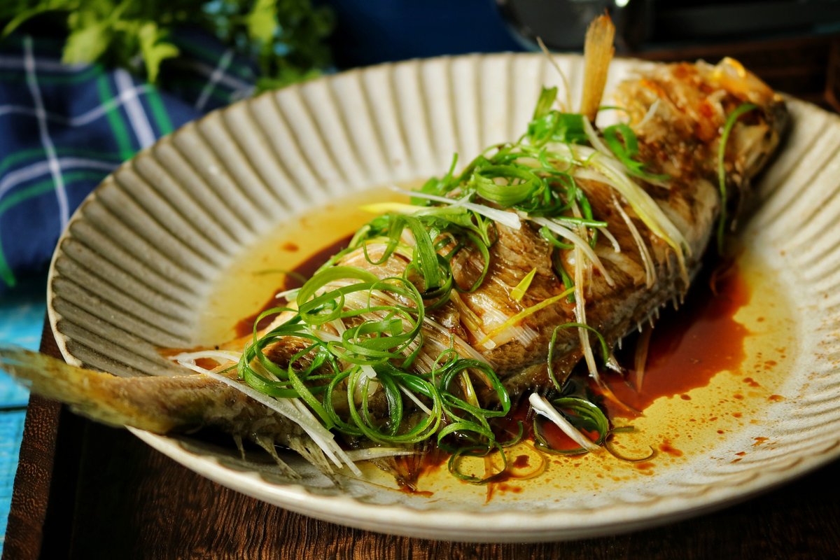 Steamed fish on a family dinner table — surrounded by other dishes, chopsticks reaching in, rice bowls nearby, the centerpiece of a Cantonese meal