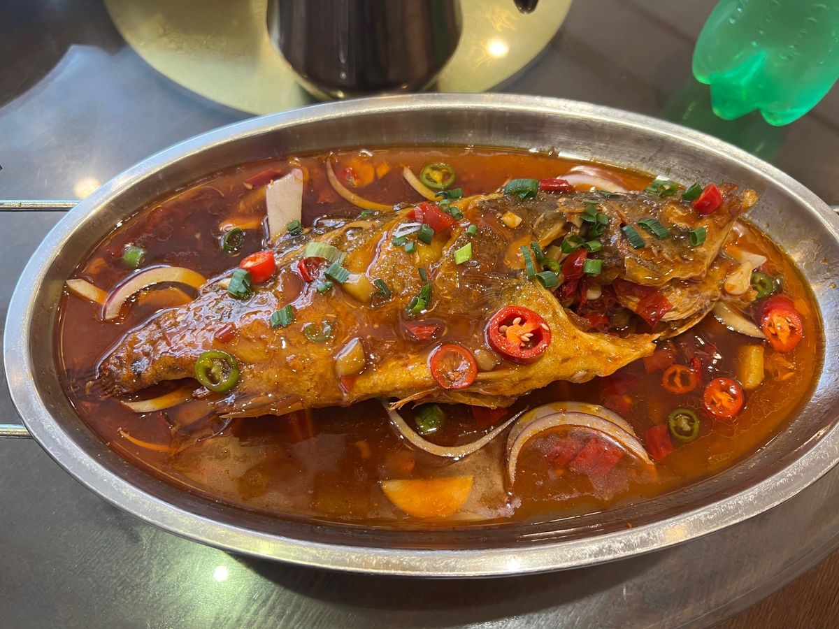 Stinky mandarin fish braised whole in a stainless pan, glossy red sauce and sliced chilli over the back, on a metal table at a small Anhui restaurant