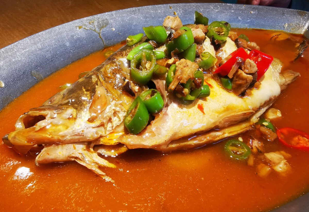 Close-up of braised stinky mandarin fish split open, the white flesh separating into firm garlic-clove flakes under a chilli sauce