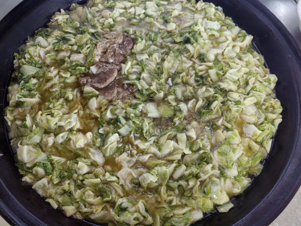 Suan cai — fermented napa cabbage stewing in a black wok with a piece of pork, glossy with oil