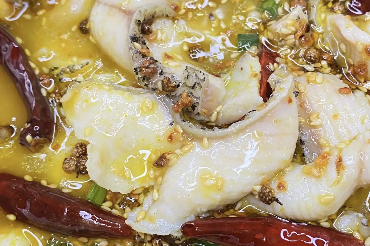 Close-up of Suan Cai Yu — a slice of fish cradling broth, pickled mustard green, and a red chili visible in the golden liquid