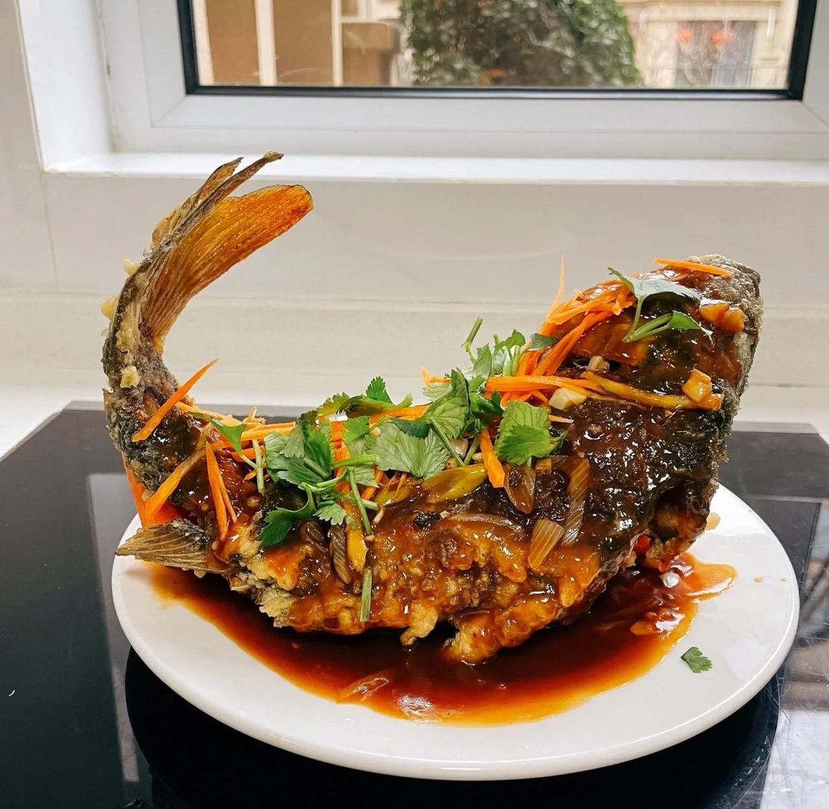 Sweet-and-sour carp at home — tail curled high, sauce just poured over the fried fish, on an oval plate by a kitchen window