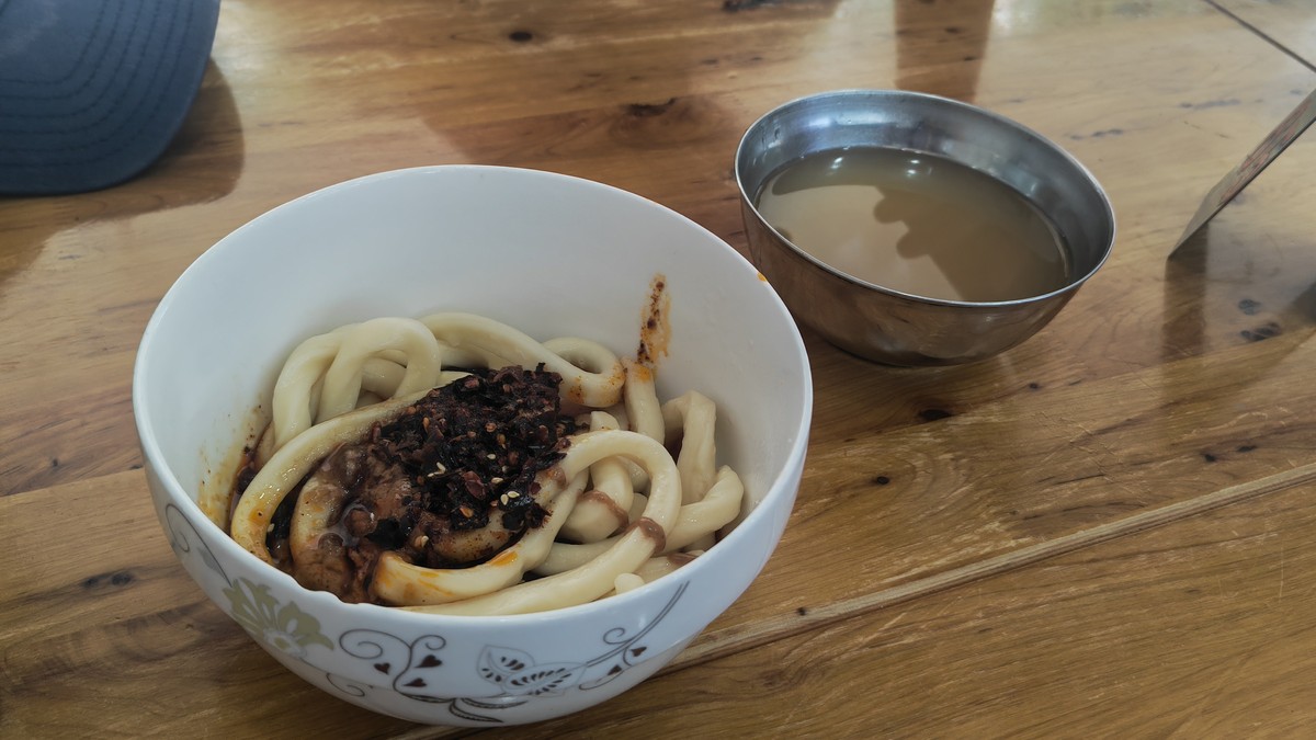 Sweet water noodles — a small white bowl of chopstick-thick noodles in dark sweet soy sauce on a worn wooden table in a Chengdu noodle shop