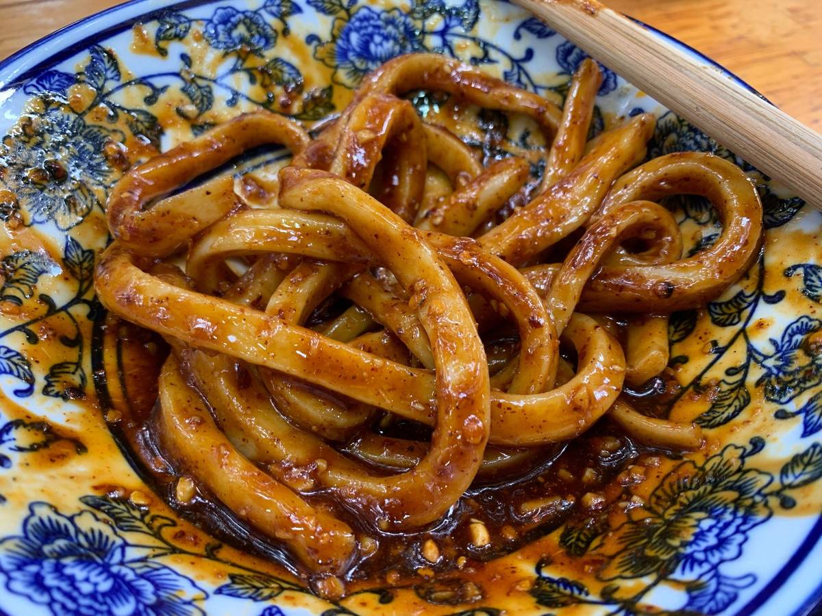 Sweet water noodles close up — thick pale noodles lifted clear of dark chilli oil sauce, the sediment and crushed peanuts visible in the bowl