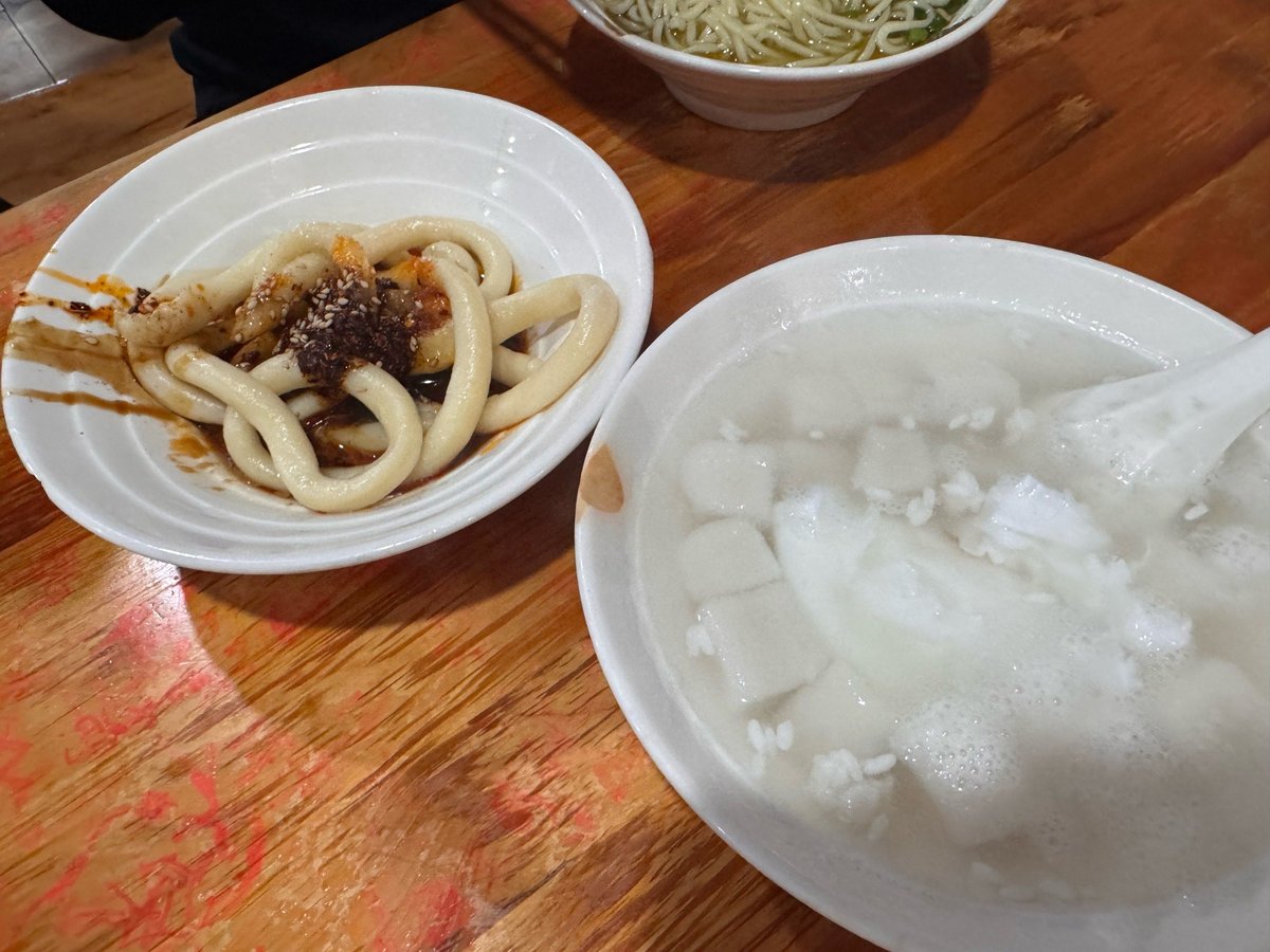 The table at a Chengdu snack shop — a plate of sweet water noodles pushed to the left, other small dishes and a bowl of sweet tofu dessert spread across the worn wooden tabletop