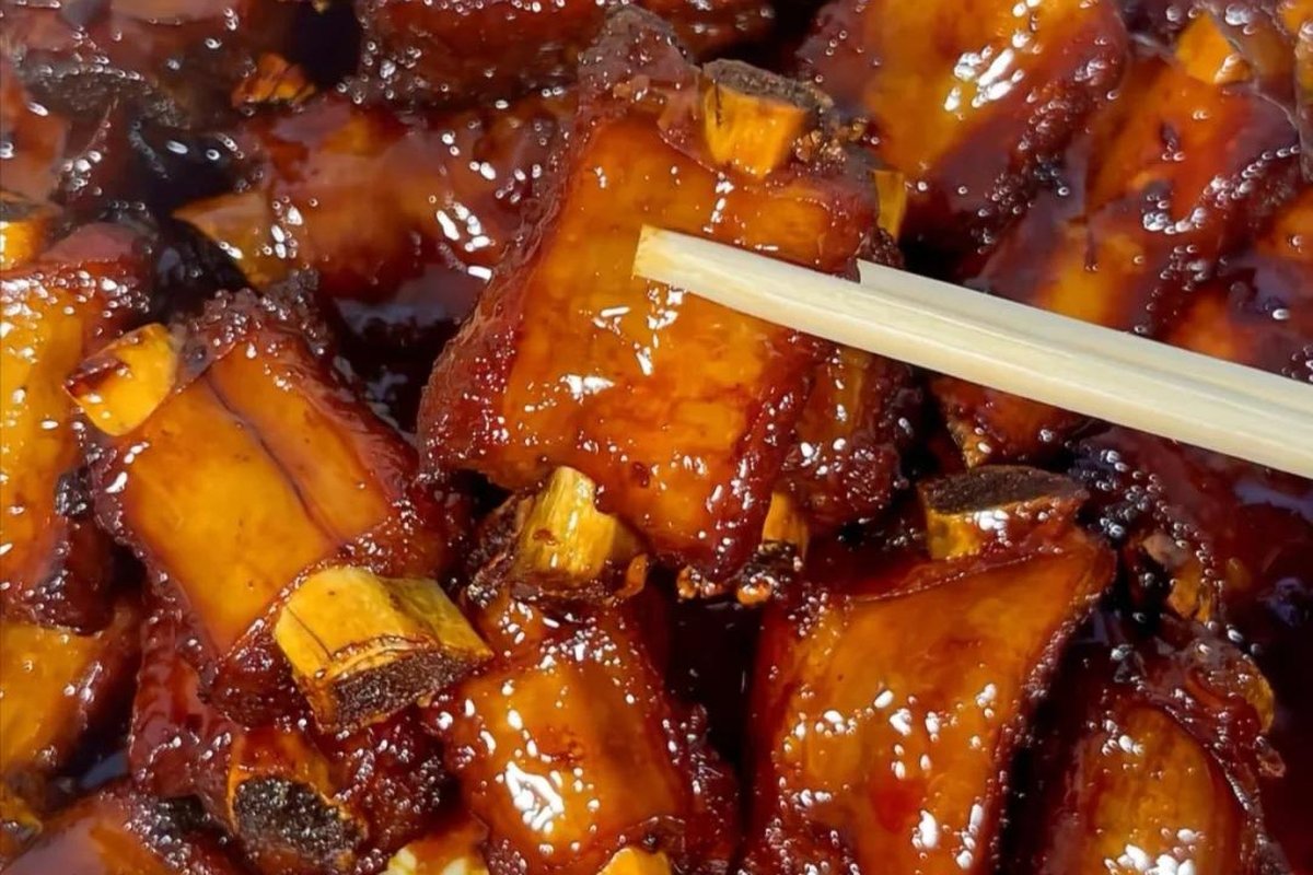 Tang Cu Pai Gu close-up — a single rib lifted by chopsticks, glossy dark amber sauce stretching, sesame seeds stuck to the glaze
