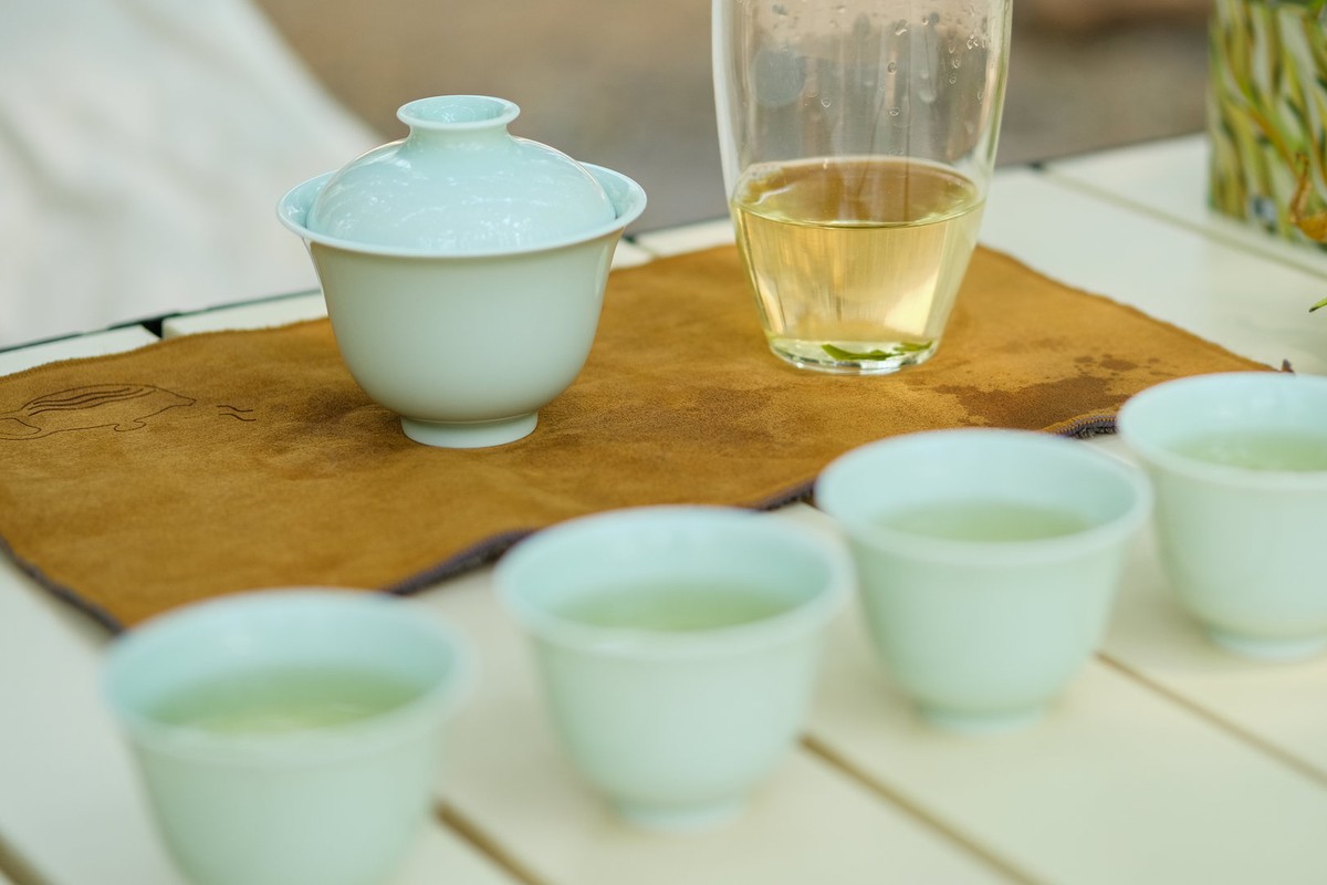 Four small white porcelain tasting cups holding pale tea in the foreground, with a gaiwan and a glass fairness pitcher behind them on a tea mat