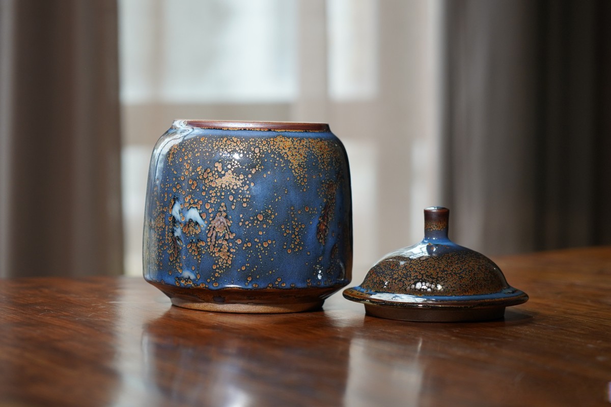 A blue tenmoku-glazed ceramic tea caddy and its lid on a polished wooden table