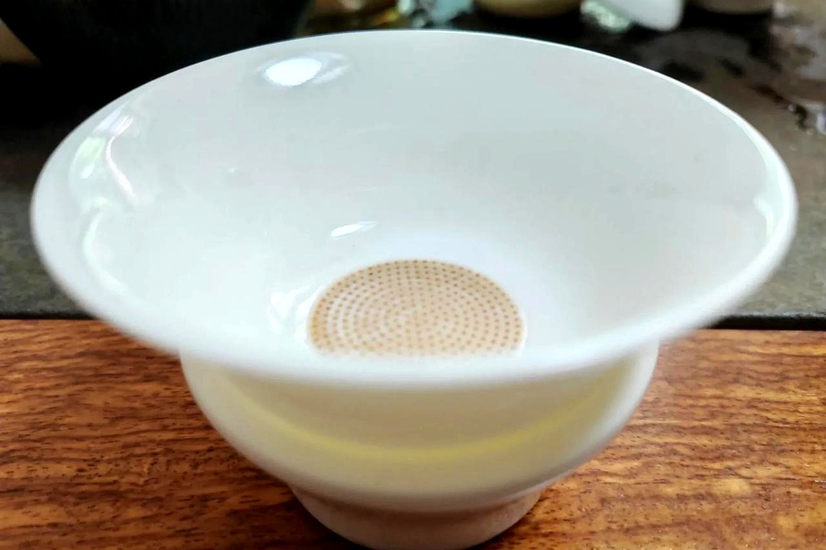 A white porcelain tea strainer with a perforated filter disc resting on a polished wooden table, blurred tea vessels in the background