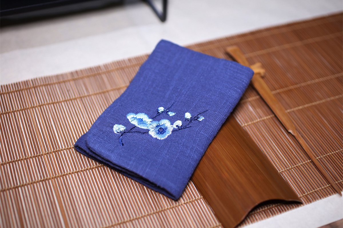A folded indigo linen tea towel with embroidered white and blue blossoms, resting on a bamboo mat beside wooden chopsticks and a chopstick rest