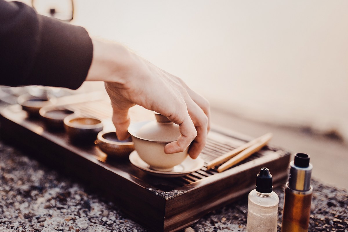 A hand lifting the lid of a white gaiwan on a dark slatted wooden tea tray, with small tasting cups and wooden tongs alongside