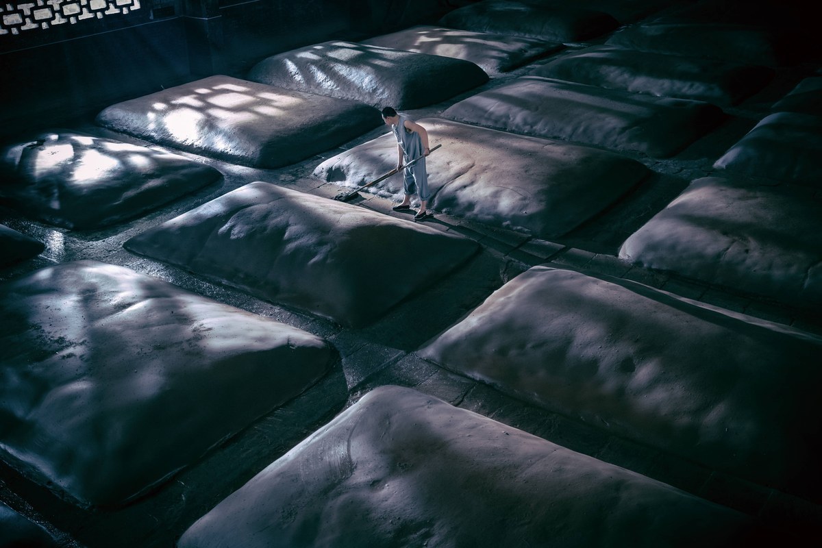 Rows of sealed fermentation pits under a single shaft of daylight in a Luzhou distillery hall, a worker tending the mud caps