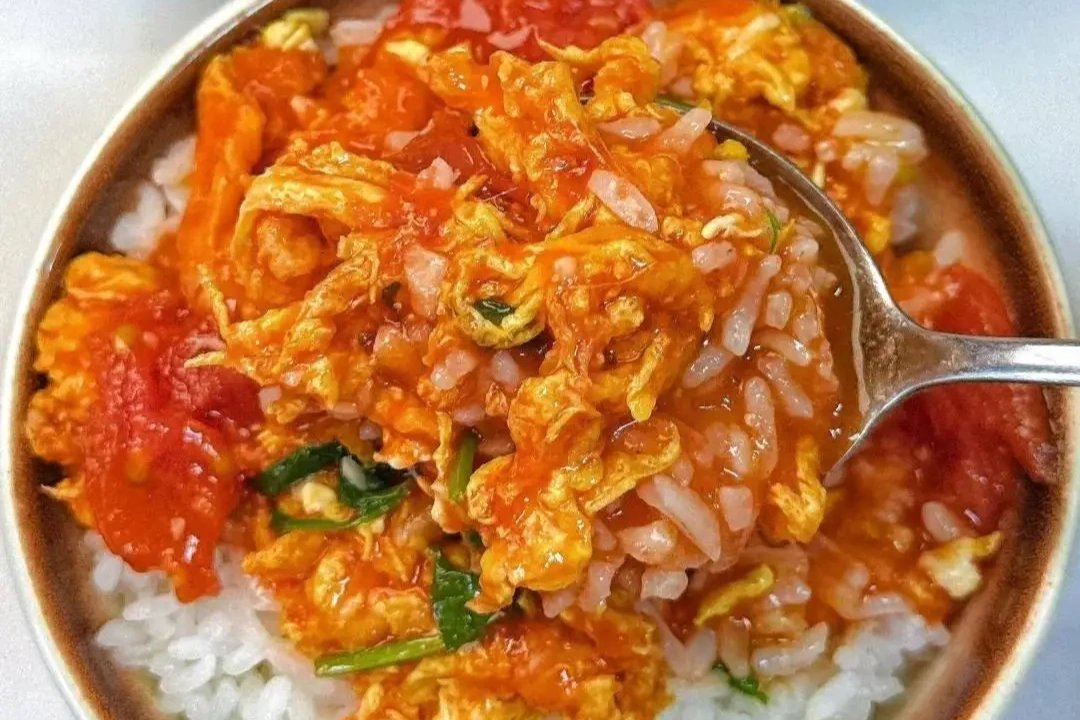 Top-down view — a spoonful of Tomato and Egg Stir-fry hovering over a bowl of rice, sauce dripping onto the white grains, steam curling upward
