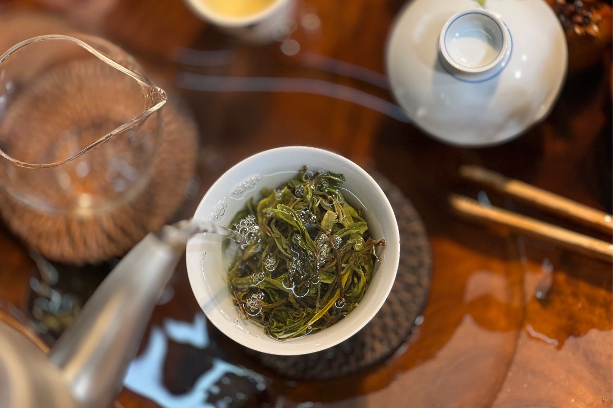 Water poured from a steel gooseneck kettle into a white porcelain bowl of green tea leaves on a wooden tea tray
