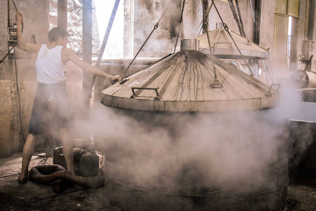 A worker in a white singlet steadying the hoist of a conical metal still lid, thick steam spilling from under it into an old distillery hall