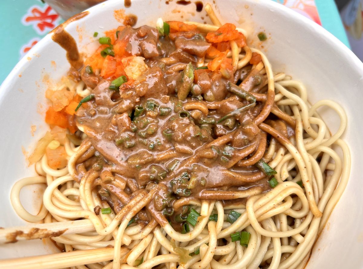 Wuhan hot-dry noodles — thick pale-yellow wheat noodles under a dense dollop of dark sesame paste, chopped scallion, diced pickled radish and chili oil, disposable chopsticks resting on the rim of the bowl