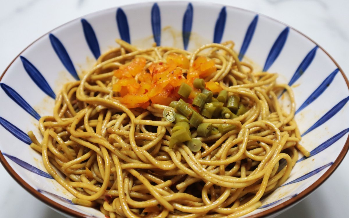 Hot-dry noodle close-up — dry-tossed yellow wheat noodles glossed with sesame paste and chili oil, topped with diced orange pickled radish and sliced scallion in a blue-and-white bowl, no broth in sight