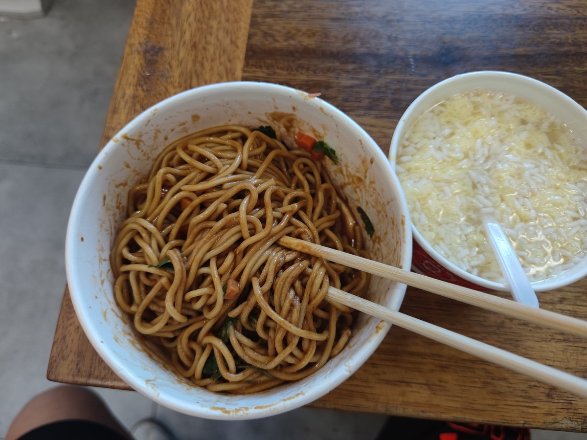 A Wuhan breakfast on a worn wooden table — a paper bowl of sauce-tossed noodles with disposable chopsticks laid across the rim, a bowl of rice in thin broth alongside