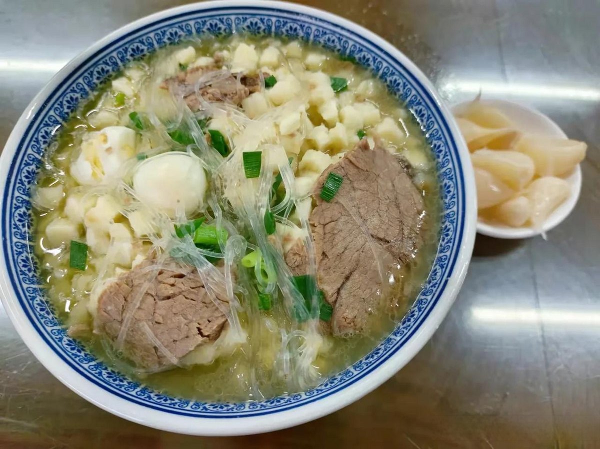 Yangrou Paomo — a deep blue-rimmed bowl of lamb broth with torn bread, glass noodles and sliced mutton, sweet pickled garlic alongside